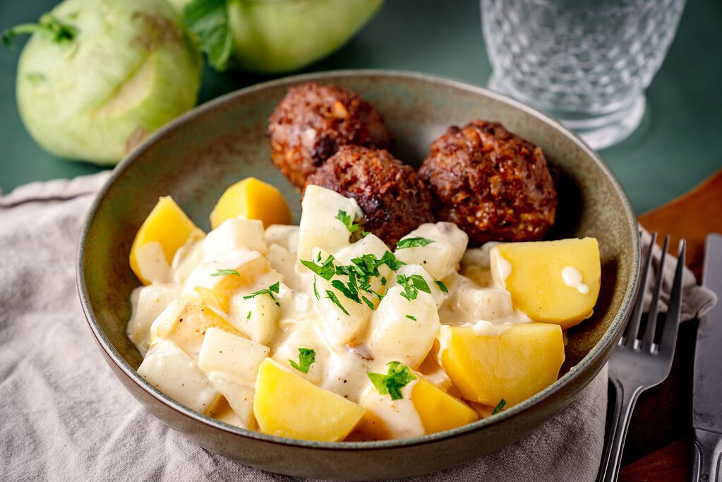 Rahm Kohlrabi mit Salzkartoffeln und Frikadellen auf einem grünen Teller auf grünem Untergrund.