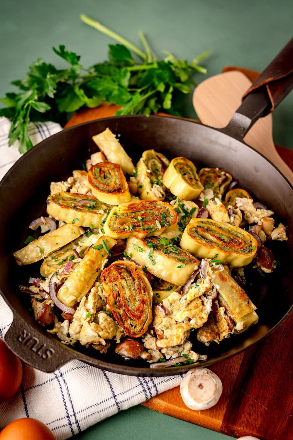Schnelle Maultaschenpfanne in einer gusseisernen Pfanne mit Ei, Zwiebeln und Champignons sowie Kräutern. Serviert auf einem Holzbrett mit grünem Untergrund