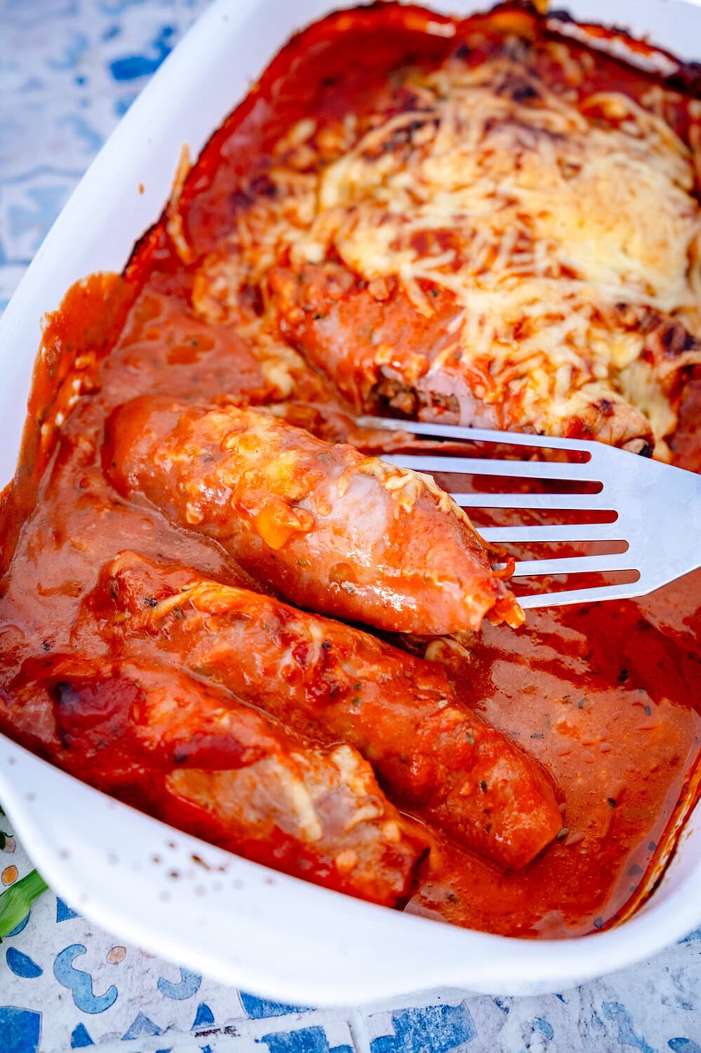 Überbackene Schinken-Hackröllchen in einer Auflaufform mit einem Pfannenwender, die gerade aus der Form geholt werden. Rote Tomaten-Sahne-Sauce und geschmolzener Käse sind zu sehen.