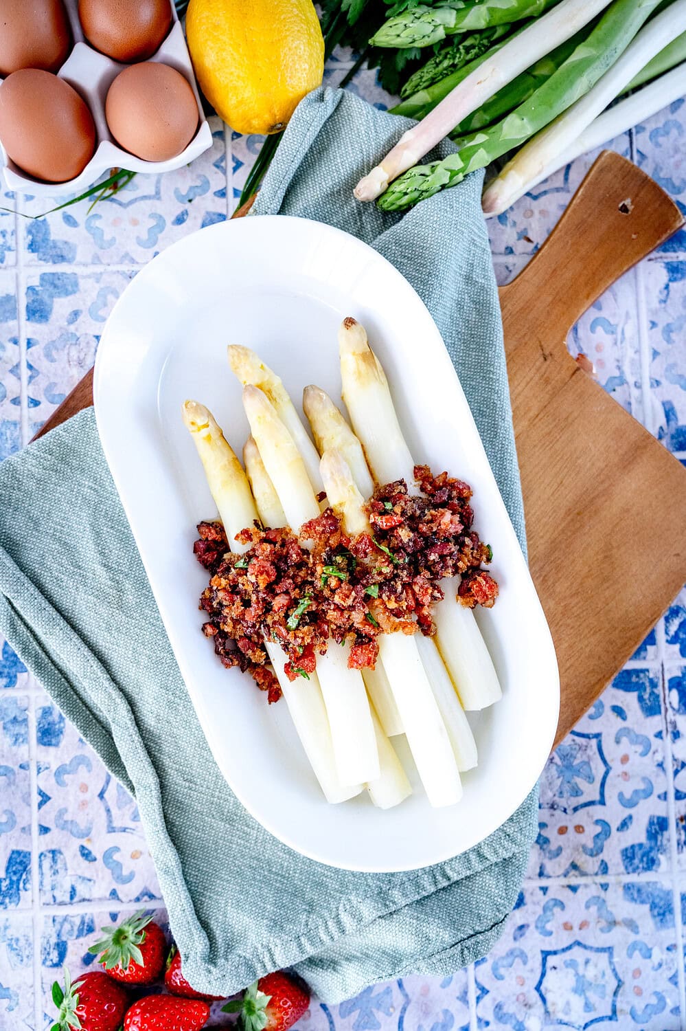 Weißer Teller mit Spargel und Specksauce auf einem Holzbrett umgeben von Spargel und Erdbeeren.