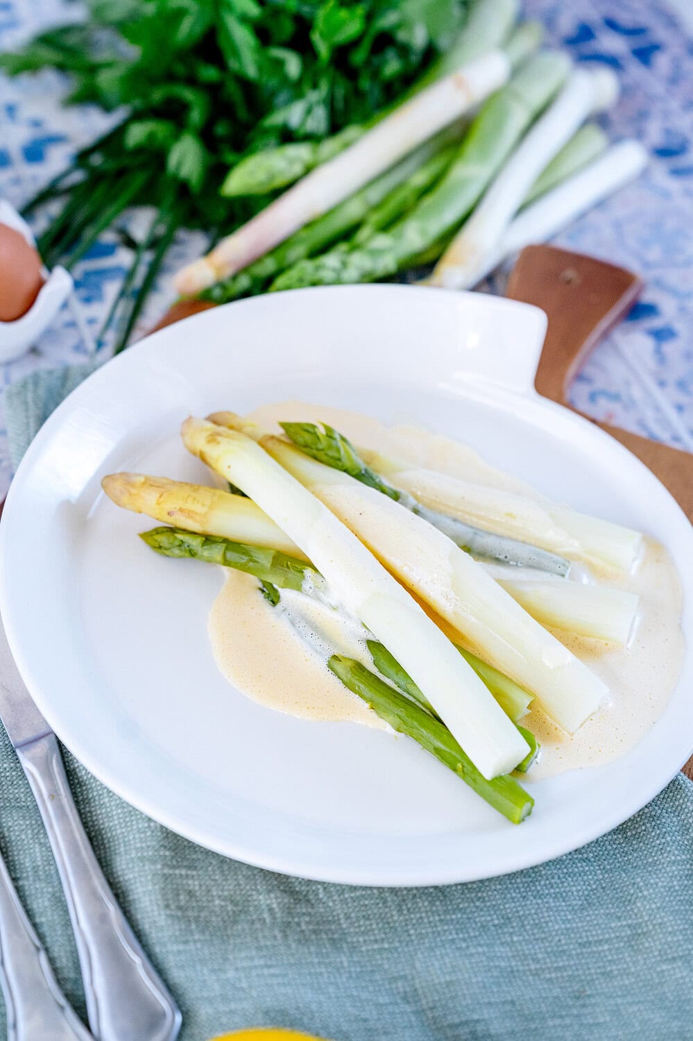weißer und grüner Spargel auf einem weißen Teller mit einer Blitz Hollandaise. Im Hintergrund sind Kräuter und frischer Spargel zu sehen.