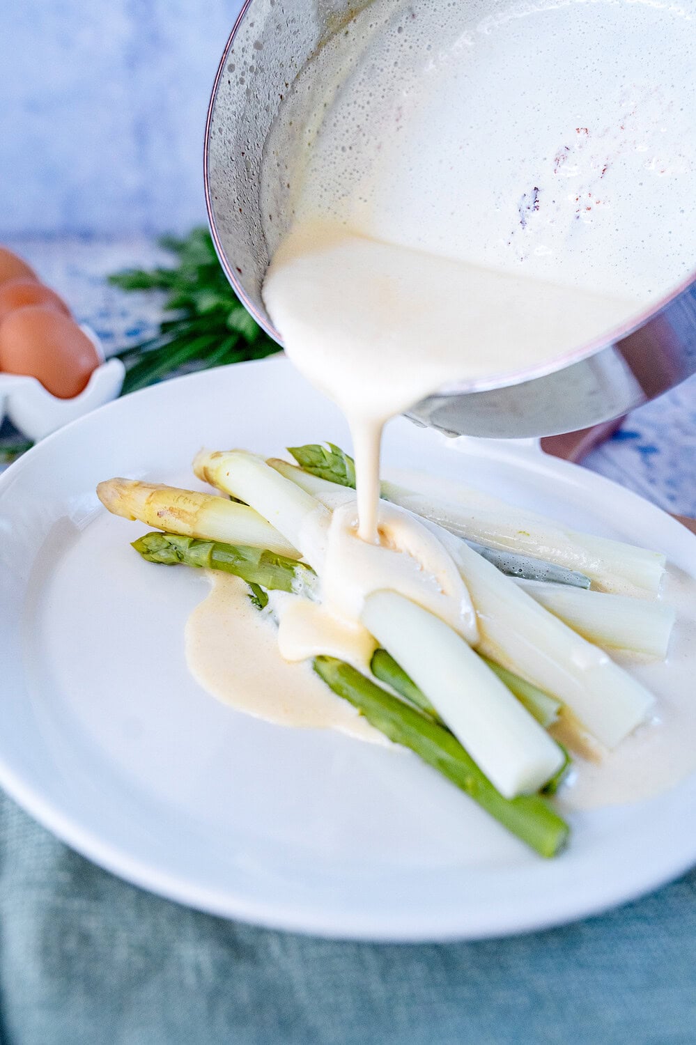 Blitz Hollandaise, die gerade aus einem Topf auf den Teller mit grünem und weißem Spargel gegossen wird.